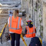 Dwoje archeologów w pomarańczowych kamizelkach wykonuje pomiary geodezyjne przy kamiennym murze.