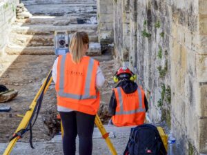 Dwoje archeologów w pomarańczowych kamizelkach wykonuje pomiary geodezyjne przy kamiennym murze.
