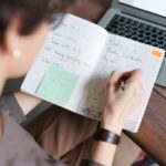 Osoba zapisująca odręczne notatki w otwartym notesie obok laptopa.