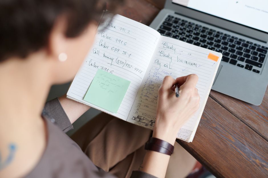 Osoba zapisująca odręczne notatki w otwartym notesie obok laptopa.