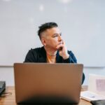 Mężczyzna siedzący przy biurku z laptopem i notatnikiem przed białą tablicą.