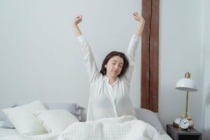 Kobieta w białej koszuli przeciąga się siedząc w łóżku po przebudzeniu