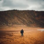 Osoba w kurtce idzie boso po piaszczystej plaży w pochmurny dzień