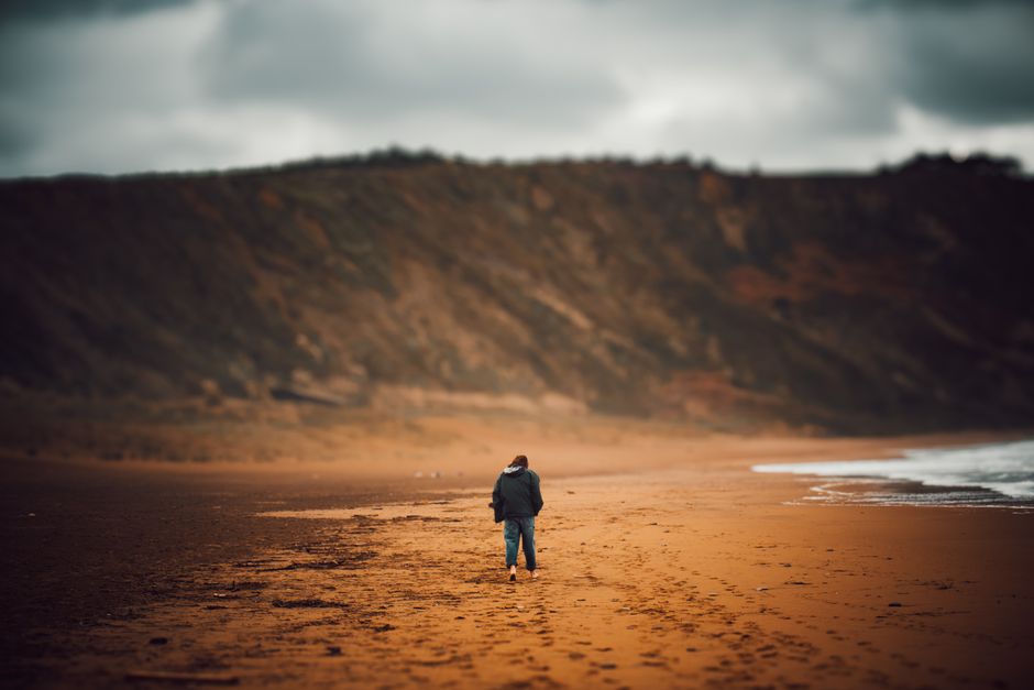 Osoba w kurtce idzie boso po piaszczystej plaży w pochmurny dzień