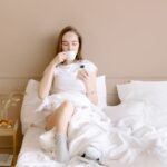 Kobieta w łóżku pije kawę i przegląda telefon przy zapalonej świecy.