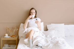 Kobieta w łóżku pije kawę i przegląda telefon przy zapalonej świecy.