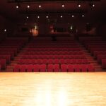 Pusta sala teatralna z rzędami czerwonych foteli i oświetloną drewnianą sceną.