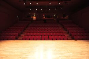 Pusta sala teatralna z rzędami czerwonych foteli i oświetloną drewnianą sceną.