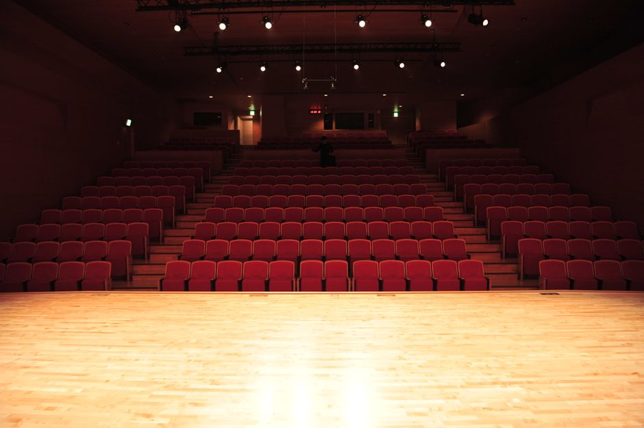 Pusta sala teatralna z rzędami czerwonych foteli i oświetloną drewnianą sceną.