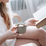 Kobieta w białej koszulce trzymająca kubek herbaty i otwartą książkę