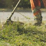 Pracownik w pomarańczowych spodniach przycina trawę za pomocą podkaszarki mechanicznej