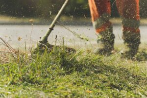 Pracownik w pomarańczowych spodniach przycina trawę za pomocą podkaszarki mechanicznej