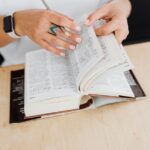 Dłonie z ołówkiem przeglądające gruby słownik leżący na drewnianym stole.