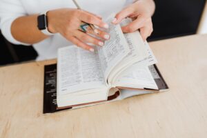 Dłonie z ołówkiem przeglądające gruby słownik leżący na drewnianym stole.